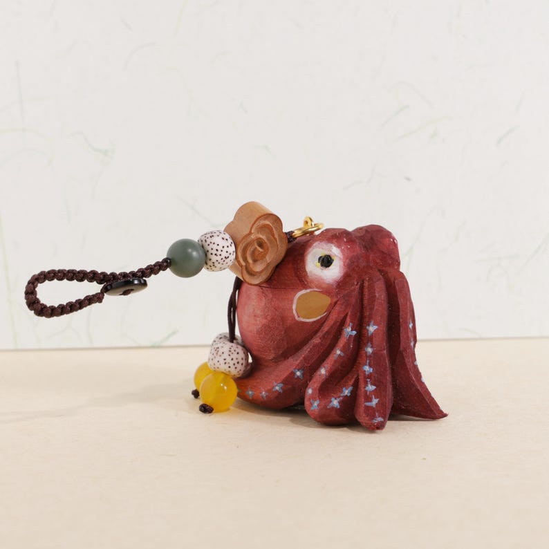 May include: A hand-carved, red octopus keychain with a brown braided loop. The octopus has white star accents and a yellow bead. The keychain also features wooden and speckled beads.