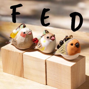 May include: Three small carved bird figurines labeled F, E, and D. The first is beige with red cheek patches, the second is white with yellow and brown accents, and the third is orange and brown. Each bird features a small beaded charm attachment.