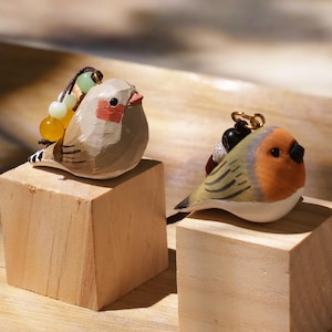 May include: Two hand-carved wooden bird figurines. The left bird is light brown with a red cheek patch and a string of yellow and green beads. The right bird has an orange face, grey wings, and a black beak, featuring a dark cord with black beads.