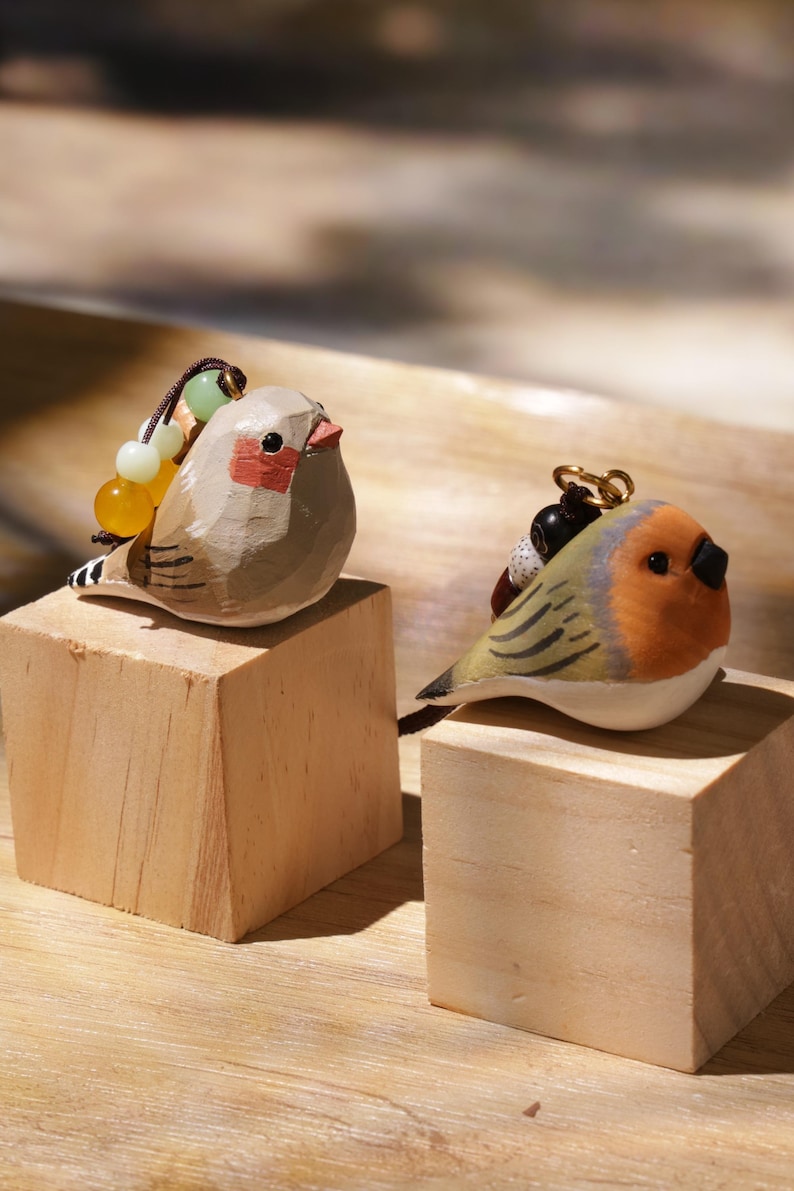May include: Two hand-carved wooden bird figurines. The left bird is light brown with a red cheek patch and a string of yellow and green beads. The right bird has an orange face, grey wings, and a black beak, featuring a dark cord with black beads.