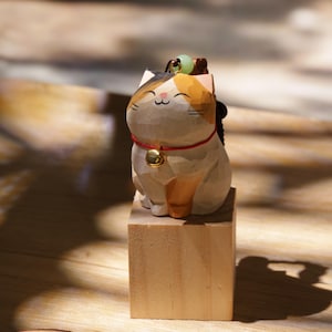 May include: A wooden cat figurine with a smiling face, wearing a red collar with a gold bell. The cat has a black, orange, and white color scheme and sits atop a small wooden block. A small green bead sits on its head.