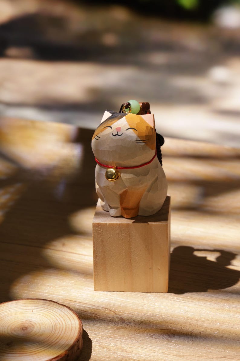 May include: A wooden cat figurine with a smiling face, wearing a red collar with a gold bell. The cat has a black, orange, and white color scheme and sits atop a small wooden block. A small green bead sits on its head.