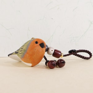 May include: A ceramic bird figurine with orange, green, and white coloring. The bird has a black beak and eyes. Attached is a brown cord with decorative beads. The figurine is a charming decorative item.