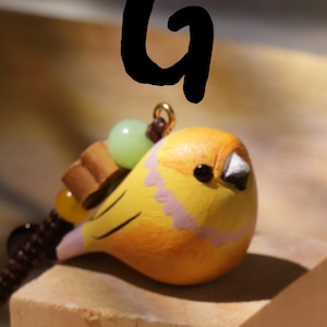 May include: A yellow bird figurine charm with a black eye and white beak, featuring a pink scalloped pattern on its chest. The charm is attached to a brown cord with a light green bead, a small brown wooden piece, and a yellow bead, marked with a black letter G.