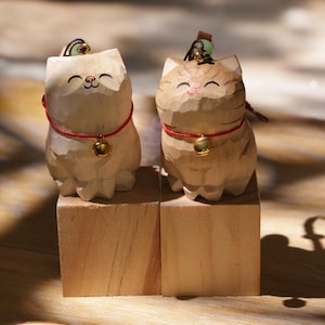 May include: Two carved wooden cat figurines. One is light beige, the other is tan with brown stripes. Both have red collars with gold bells and small green accents on their heads. They sit on wooden blocks.