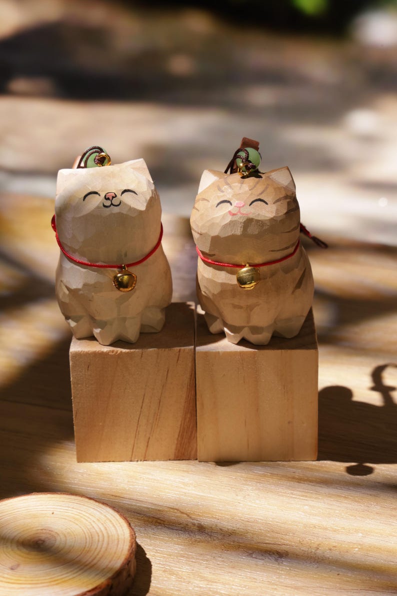 May include: Two carved wooden cat figurines. One is light beige, the other is tan with brown stripes. Both have red collars with gold bells and small green accents on their heads. They sit on wooden blocks.