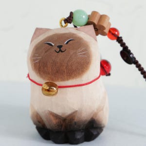 May include: A wooden cat figurine with a smiling face, brown accents, and a gold bell on a red cord. The cat is part of a beaded charm with wooden and red beads.