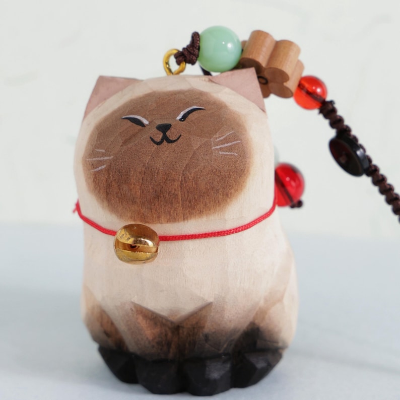May include: A wooden cat figurine with a smiling face, brown accents, and a gold bell on a red cord. The cat is part of a beaded charm with wooden and red beads.