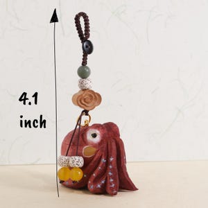 May include: A handcrafted, red wooden octopus charm, approximately 4.1 inches in length, with a decorative cord and beads. The charm features a detailed octopus design with white accents and is adorned with various beads in shades of brown, green, and yellow.