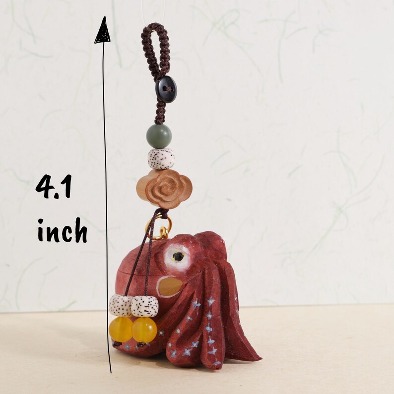 May include: A handcrafted, red wooden octopus charm, approximately 4.1 inches in length, with a decorative cord and beads. The charm features a detailed octopus design with white accents and is adorned with various beads in shades of brown, green, and yellow.