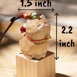 May include: A wooden cat figurine, 2.2 inches tall and 1.5 inches wide, with a carved design. The cat has a red collar and a small gold bell. It is mounted on a small wooden block and has a decorative cord with beads.