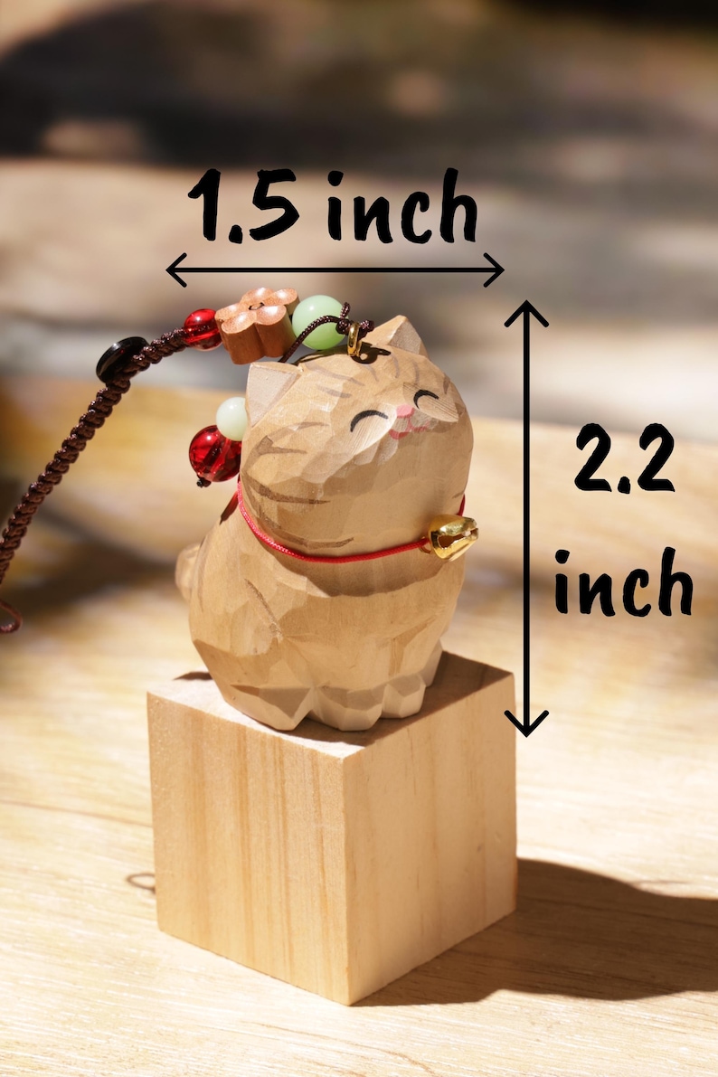 May include: A wooden cat figurine, 2.2 inches tall and 1.5 inches wide, with a carved design. The cat has a red collar and a small gold bell. It is mounted on a small wooden block and has a decorative cord with beads.