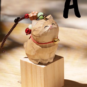 May include: A carved wooden cat figurine with a red collar and a gold bell, sitting on a small wooden block. A beaded charm with red, white, and green beads and a flower charm is attached. The letter "A" is in the background.