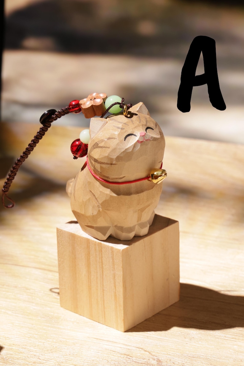 May include: A carved wooden cat figurine with a red collar and a gold bell, sitting on a small wooden block. A beaded charm with red, white, and green beads and a flower charm is attached. The letter "A" is in the background.
