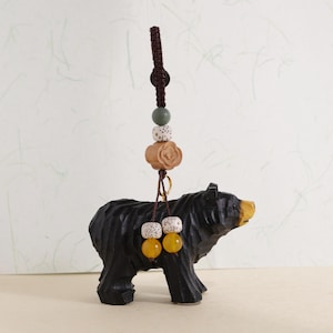 Könnte beinhalten: Eine schwarze, geschnitzte Holz-Bärenfigur mit gelber Nase und einem dekorativen Perlenanhänger. Der Anhänger hat eine braune Kordel, grüne, weiße und gelbe Perlen sowie eine Holzblume.