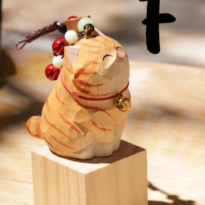May include: A carved wooden cat figurine with orange and brown stripes, sitting on a small wooden block. The cat has a red collar with a gold bell and is adorned with decorative beads. The letter "F" is in the background.