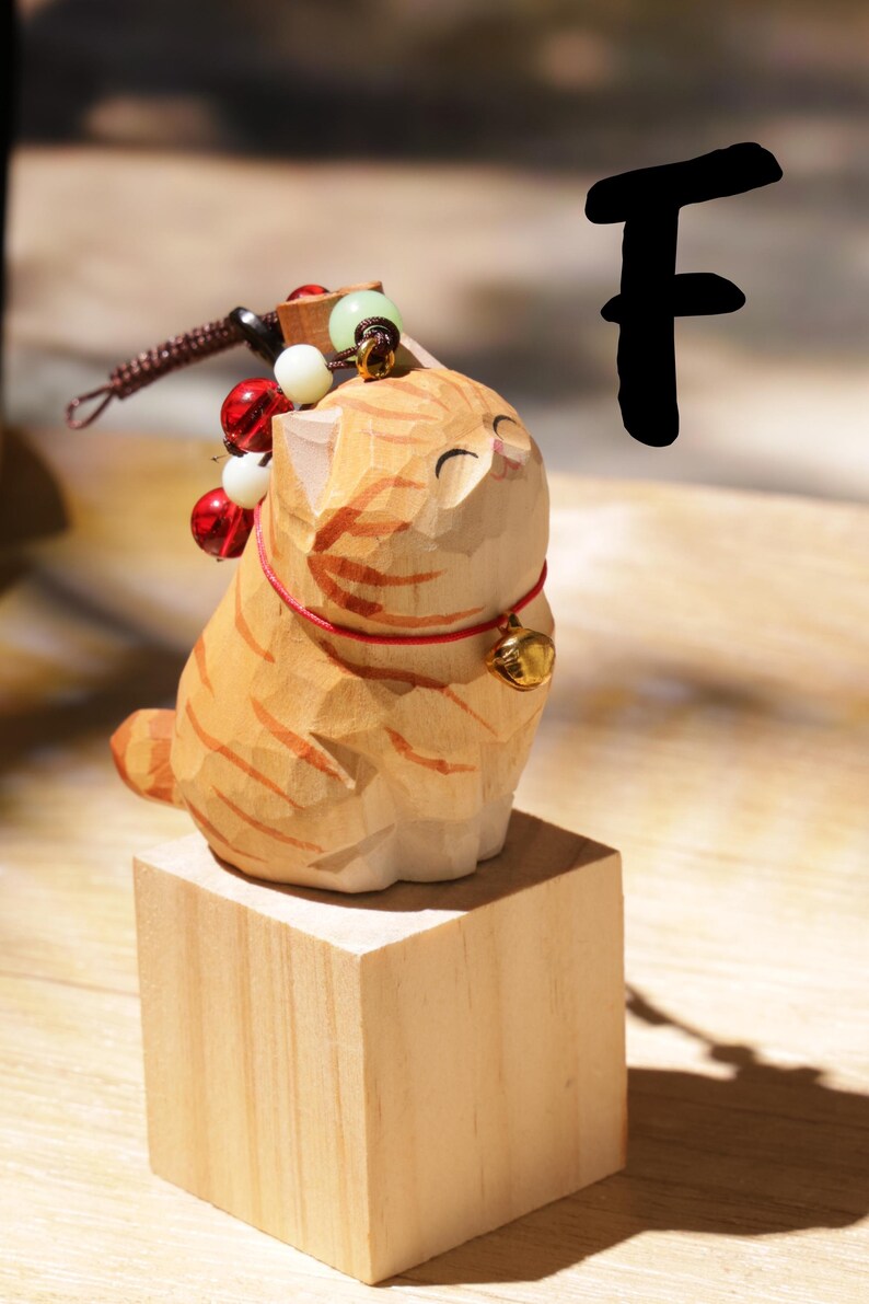 May include: A carved wooden cat figurine with orange and brown stripes, sitting on a small wooden block. The cat has a red collar with a gold bell and is adorned with decorative beads. The letter "F" is in the background.
