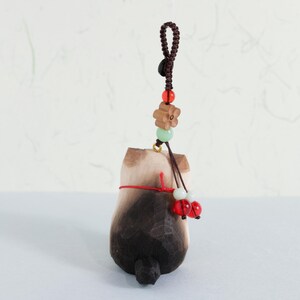 May include: A wooden cat figurine with a dark brown and light brown finish, hanging from a brown cord with decorative beads. The cat has a red string tied around its middle and two red beads hanging from the bottom.