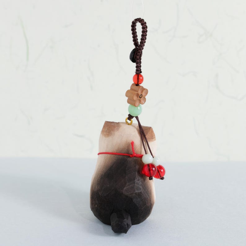 May include: A wooden cat figurine with a dark brown and light brown finish, hanging from a brown cord with decorative beads. The cat has a red string tied around its middle and two red beads hanging from the bottom.