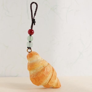 May include: A wooden croissant charm, intricately carved and suspended from a brown cord with red and green beads. The charm is light brown, and the cord features a small clasp.