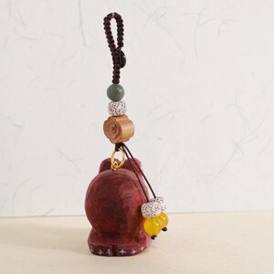 May include: A carved wooden figurine in shades of red-brown, with a decorative cord and bead accent. The figurine has a detailed, textured surface and is suspended from a dark brown cord with a knot. Additional beads include a green, speckled white, and yellow beads.