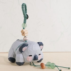 May include: A hand-carved wooden koala bear figurine in gray, with pink cheeks and black accents. It hangs from a green cord with wooden beads and a flower charm. The koala is lying down, with a sweet expression.