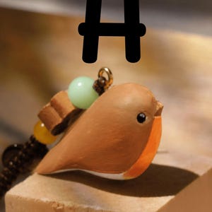 May include: A brown and orange bird figurine pendant featuring a small black eye, a white underbelly, and a dark brown cord attachment adorned with a light green bead, a yellow bead, and a small brown wooden accent piece, marked with a black letter H.