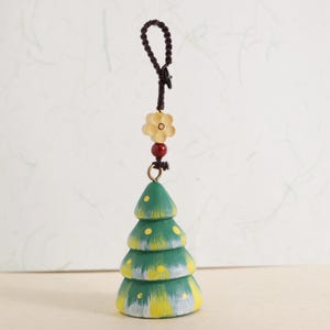 Op de afbeelding: Een groene, gelaagde kerstboomversiering met gele accenten. De versiering wordt opgehangen door een bruin koord met een bloem- en kralenversiering. De boom heeft een rustieke, handgeschilderde uitstraling.