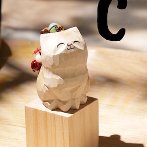 May include: A carved wooden cat figurine with a smiling face, standing on a small wooden block. The cat has a small accessory on its head and a red and white ornament on its side. A large black letter "C" is in the background.