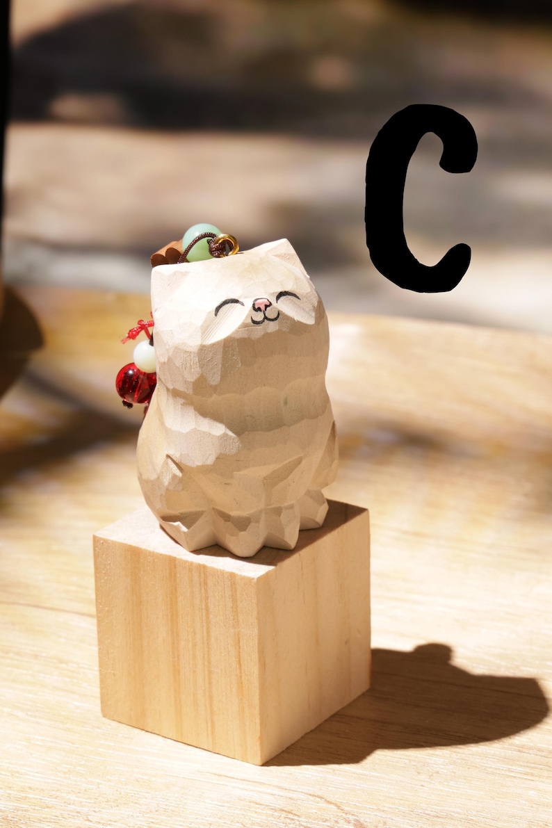 May include: A carved wooden cat figurine with a smiling face, standing on a small wooden block. The cat has a small accessory on its head and a red and white ornament on its side. A large black letter "C" is in the background.