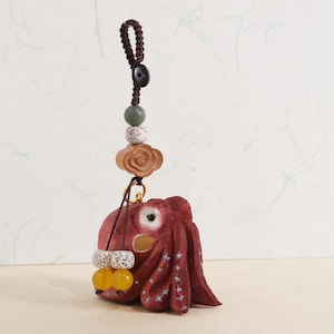 Può includere: Un ciondolo a forma di polpo in legno rosso fatto a mano con dettagli di stelle bianche, appeso a un cordino marrone con perline decorative e un fiore di legno. Il ciondolo ha perline gialle e bianche sul fondo.