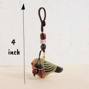May include: A handcrafted bird-shaped ornament, approximately 4 inches in length, with a brown cord and decorative beads. The bird features orange, green, and black paint, with two red-brown bead accents. The ornament is designed for hanging.