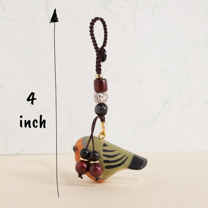 May include: A handcrafted bird-shaped ornament, approximately 4 inches in length, with a brown cord and decorative beads. The bird features orange, green, and black paint, with two red-brown bead accents. The ornament is designed for hanging.
