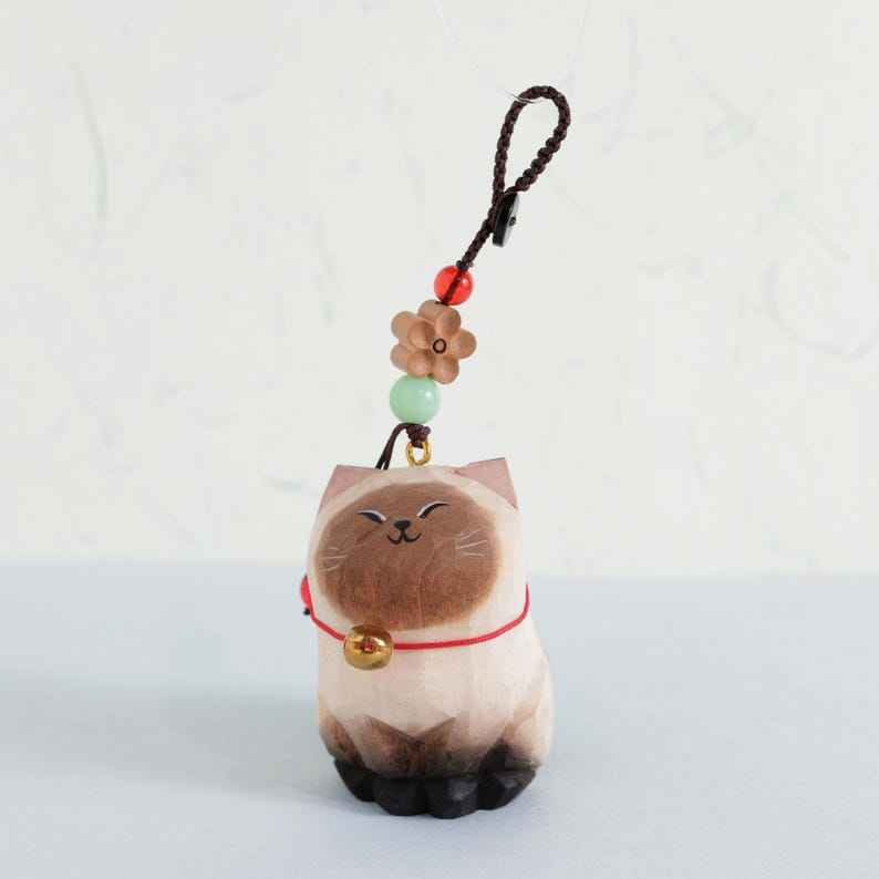 May include: A carved wooden cat figurine with a red collar and a gold bell. The cat has a smiling face and is attached to a brown cord with decorative beads and a flower. The figurine is tan, brown, and black.