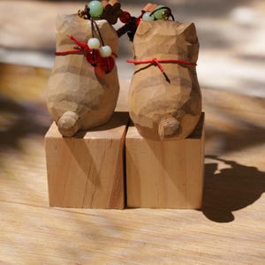 May include: Two wooden cat figurines, each carved with detailed striations, stand on separate wooden blocks. The cats have red string accents and beaded accessories. The background is a light brown wooden surface.