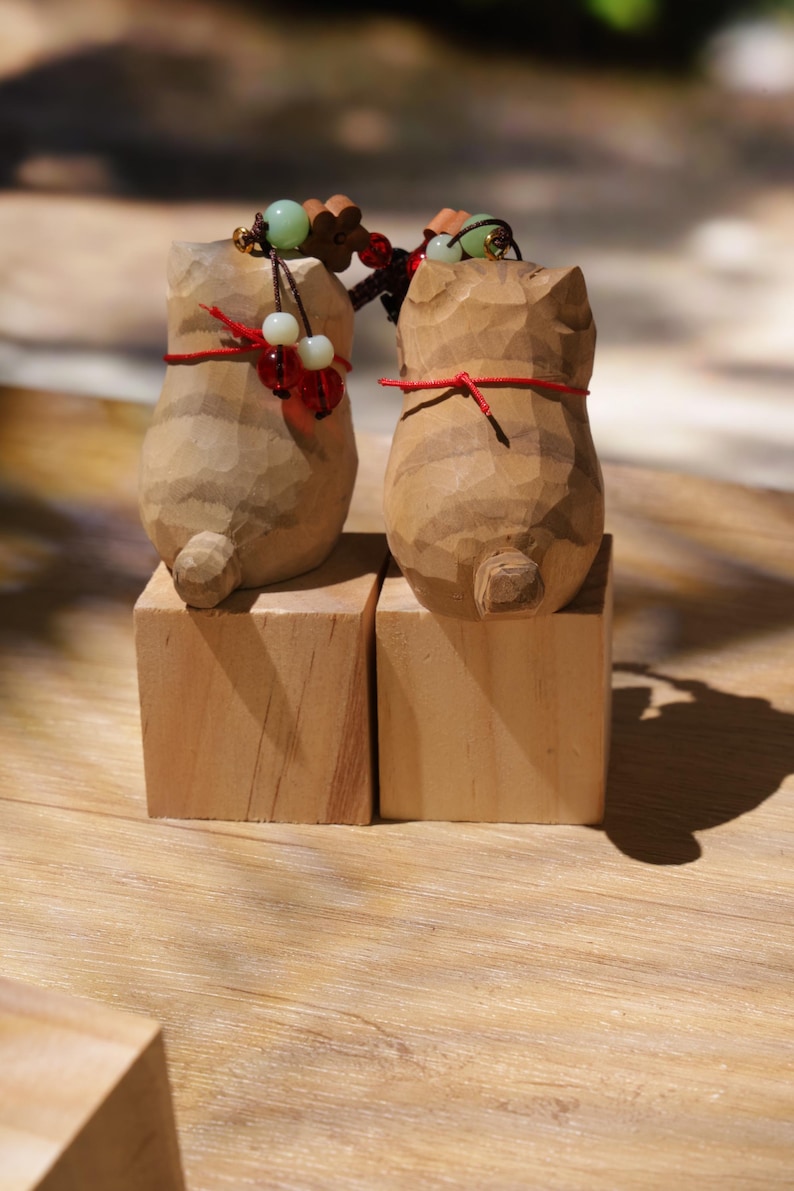 May include: Two wooden cat figurines, each carved with detailed striations, stand on separate wooden blocks. The cats have red string accents and beaded accessories. The background is a light brown wooden surface.