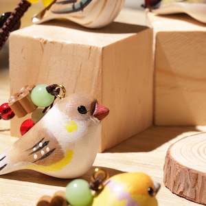 May include: Three small bird-shaped ornaments featuring hand-painted details in brown, white, and yellow. Each bird is adorned with a small brown flower-shaped bead, a translucent green bead, and a yellow bead, all attached to a dark brown cord.