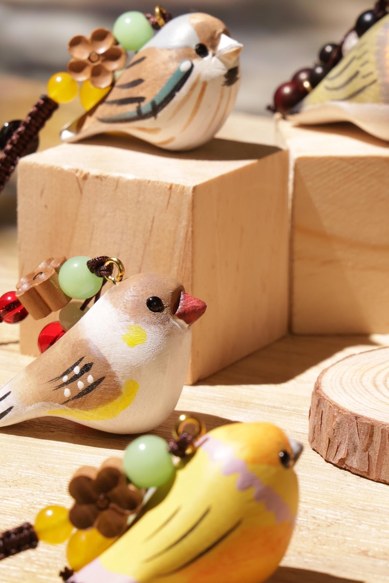 May include: Three small bird-shaped ornaments featuring hand-painted details in brown, white, and yellow. Each bird is adorned with a small brown flower-shaped bead, a translucent green bead, and a yellow bead, all attached to a dark brown cord.