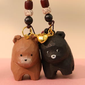 Op de afbeelding: Twee houten berenbeeldjes, één bruin en één zwart, verbonden door een gouden hartvormige bedel. De beren hebben eenvoudige gesneden kenmerken en zijn opgehangen aan een kralenkoord met donkerbruine en crèmekleurige kralen. Een charmant, handgemaakt item.