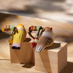 May include: Two small bird figurines, one yellow with pink accents and one brown with white markings, each featuring a dark cord adorned with small wooden flower beads and round green and red glass beads attached to the top of the bird's head.