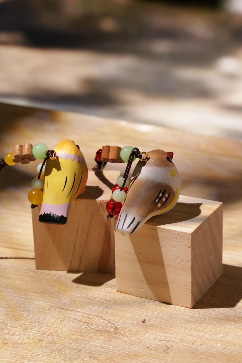 May include: Two small bird figurines, one yellow with pink accents and one brown with white markings, each featuring a dark cord adorned with small wooden flower beads and round green and red glass beads attached to the top of the bird's head.