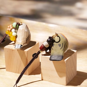 May include: Two hand-carved wooden bird charms featuring painted details, each attached to a dark brown braided cord with decorative beads. One bird is light tan with black wing markings, while the other is olive green with dark grey wing accents.