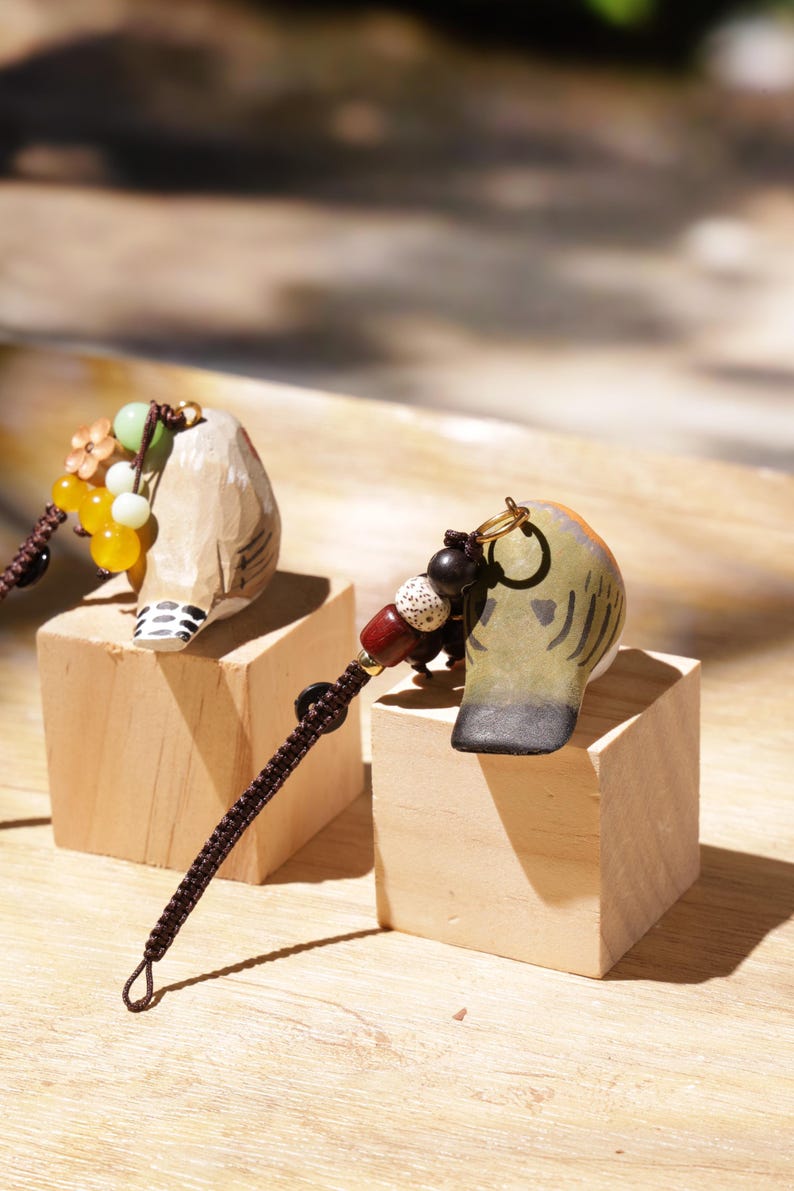 May include: Two hand-carved wooden bird charms featuring painted details, each attached to a dark brown braided cord with decorative beads. One bird is light tan with black wing markings, while the other is olive green with dark grey wing accents.