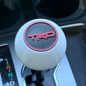 Puede incluir: Un pomo de cambio blanco con el logotipo TRD rojo y negro. El pomo redondo está montado en una palanca de cambios plateada. El fondo muestra el interior de un coche, incluido el selector de marchas.