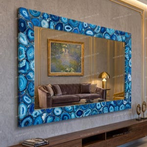 Puede incluir: Un gran espejo de pared rectangular con un marco ancho compuesto por numerosas rodajas de ágata azul pulida. El espejo refleja una habitación que contiene un sofá marrón, una lámpara de mesa dorada y un cuadro de paisaje en la pared.