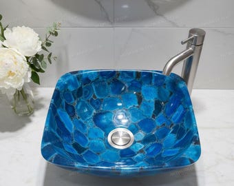 May include: A square, blue agate stone sink with a stainless steel faucet. The sink's surface is a mosaic of blue and black stones. The faucet is a modern, single-handle design. The sink is set against a white tiled wall.