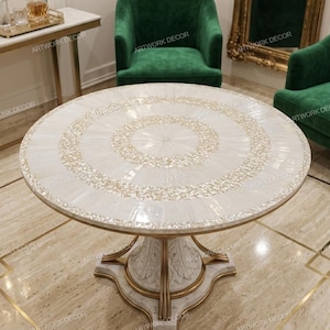 Puede incluir: Una mesa redonda con una superficie blanca y complejos patrones de nácar incrustado dispuestos en círculos concéntricos. La mesa se apoya en una base central con detalles blancos tallados y acentos de metal en tono dorado.
