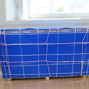 May include: A rectangular blue shipping crate wrapped in white grid lines and secured with thin white cord. The top edge features a repeating white tape strip printed with the word FRAGILE and umbrella icons. The crate rests on small wooden blocks.