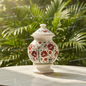 May include: A decorative white marble jar with a lid, featuring red floral designs and green detailing. The jar is set on a pedestal base. The background shows green foliage. The jar is likely around 20 cm tall.