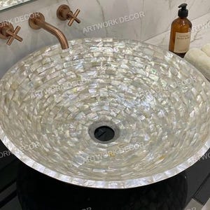 May include: A round, mosaic-tiled bathroom sink with a pearlescent finish. The sink is paired with copper-toned faucets and fixtures. A bottle of hand soap and rolled towels are visible on the countertop. The text "ARTWORK DECOR" is visible.